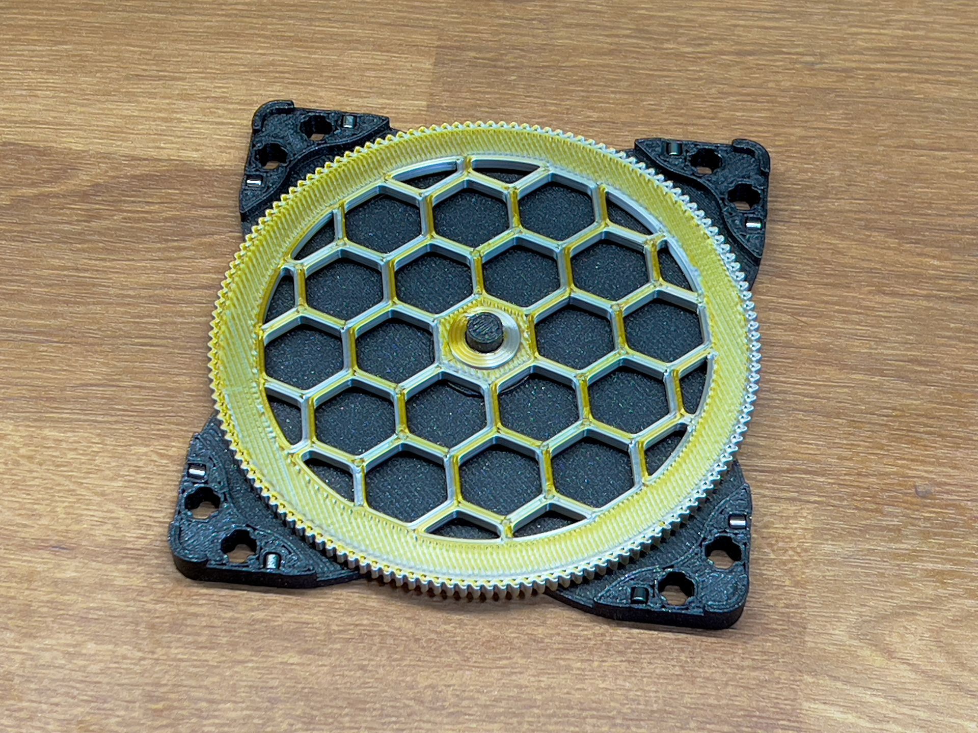 A gold, circular gear with a central honeycomb lattice pattern sits atop a black, square mounting plate on a wood surface.
