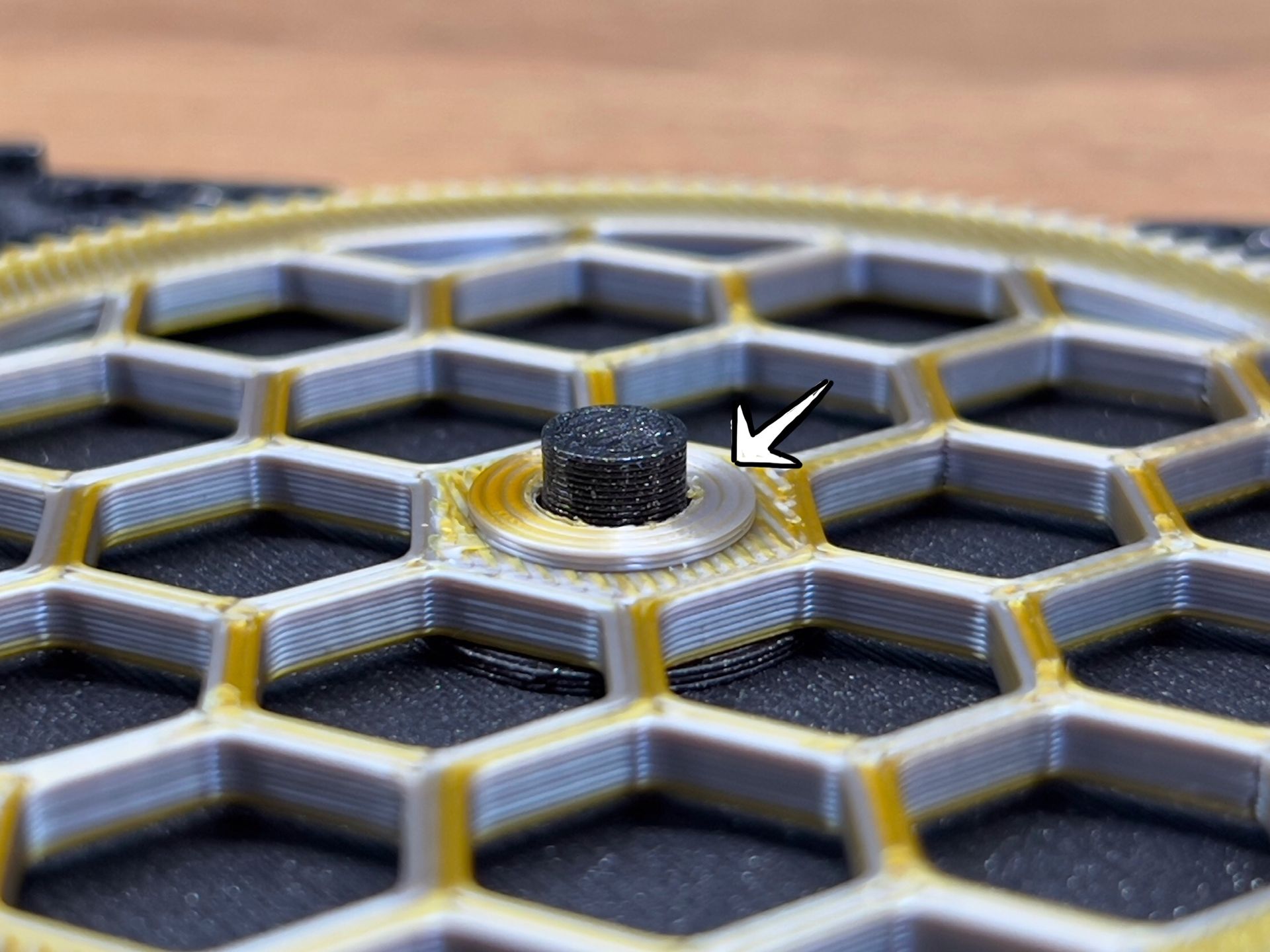 Close-up of a silver washer centered on a gray and gold 3D-printed honeycomb structure, indicated by a white arrow.