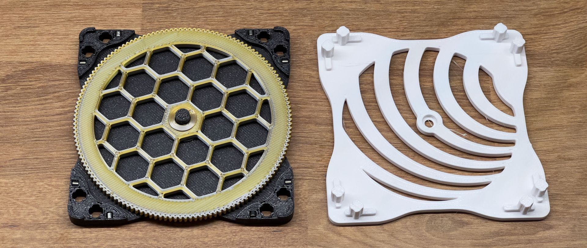 Two plastic components for 3D printing lie on a wooden surface: a square base with a gold-colored gear and a white grate.
