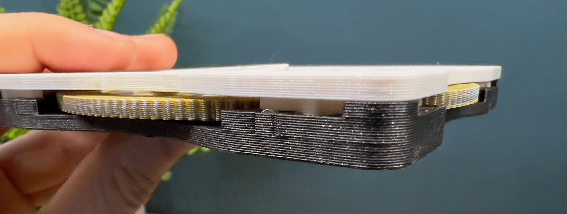 A hand holds a 3D-printed black frame containing gold gears sandwiched between white plates.