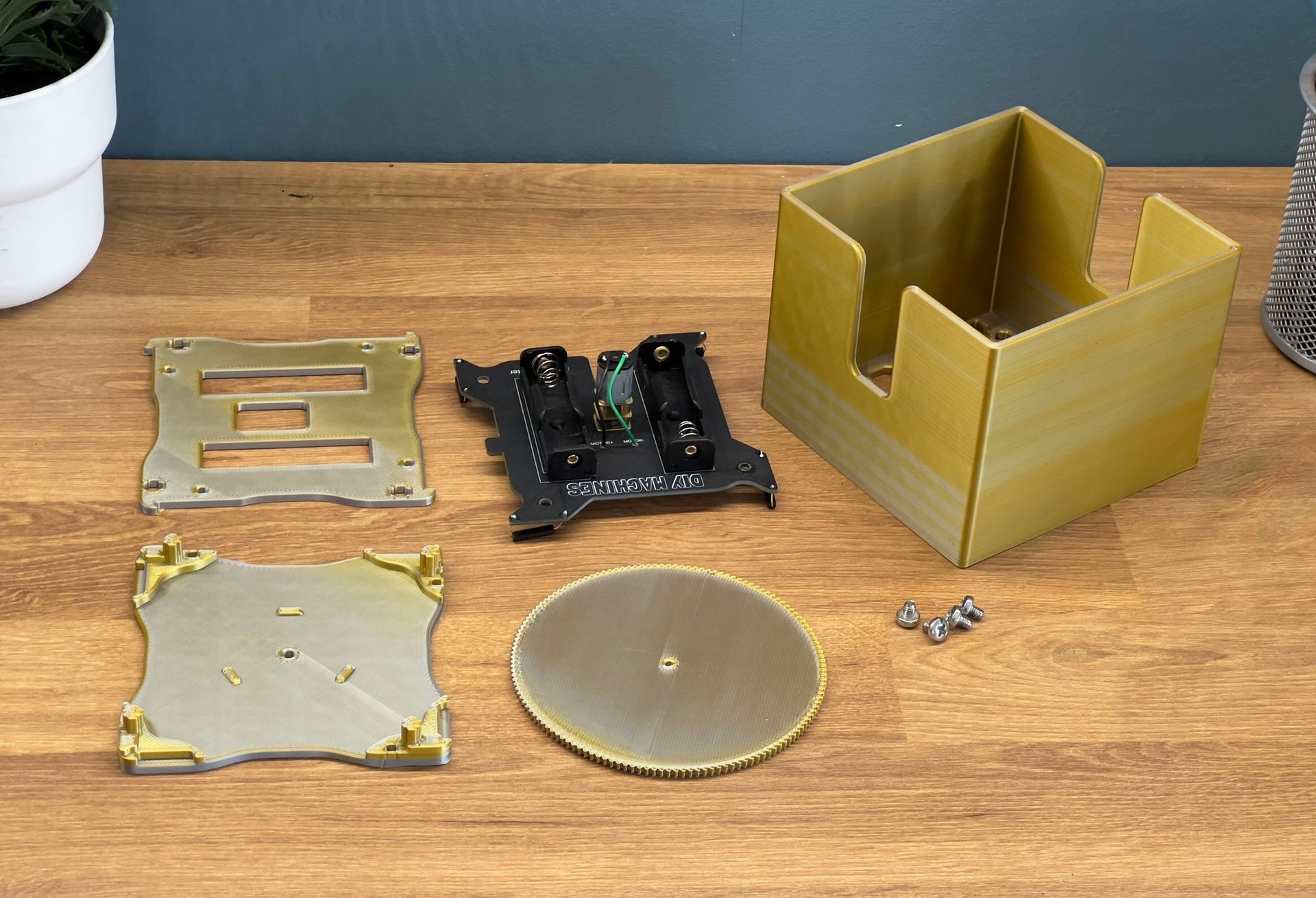 Disassembled components of a small mechanical device, including metal plates, a circuit board, and a box, on a wood table.