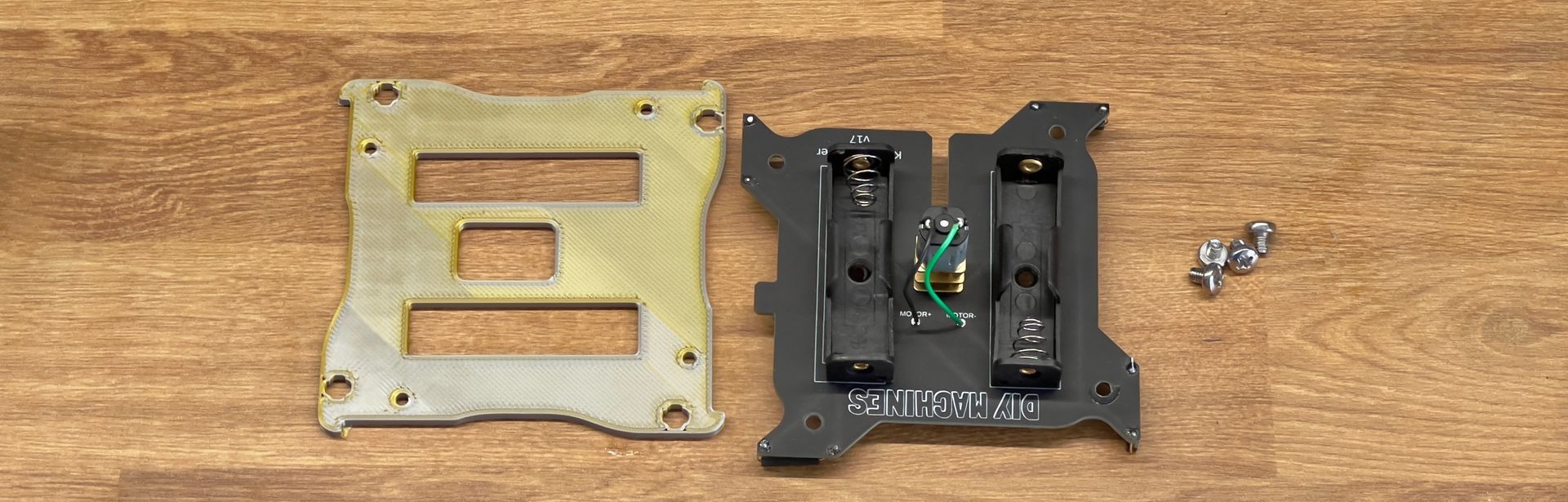 A gold metal plate, a black plastic bracket with battery compartments, and small screws laid out on a wooden surface.