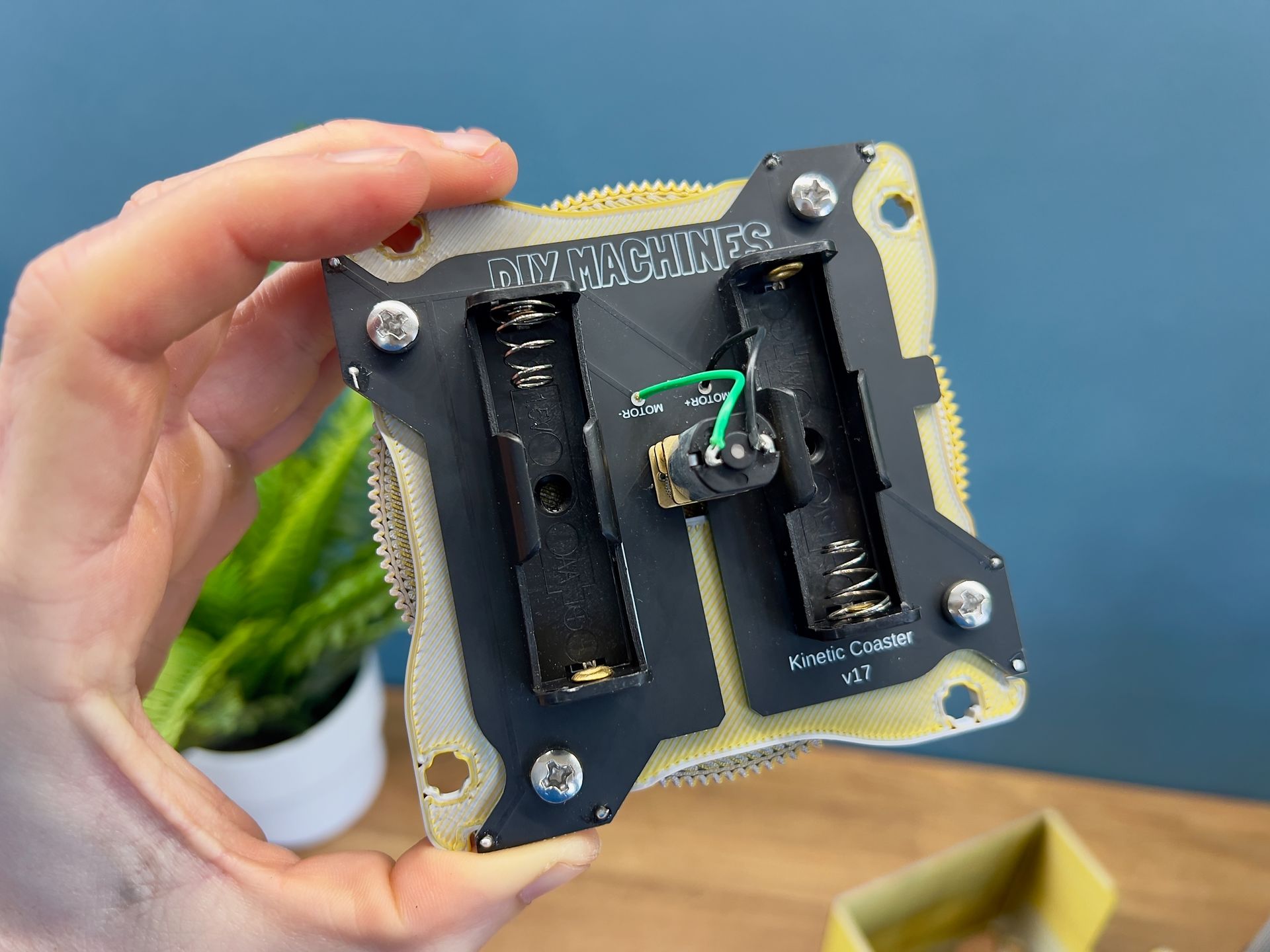 A hand holds a custom-made square electronic device featuring two battery holders and a central component with green wires.