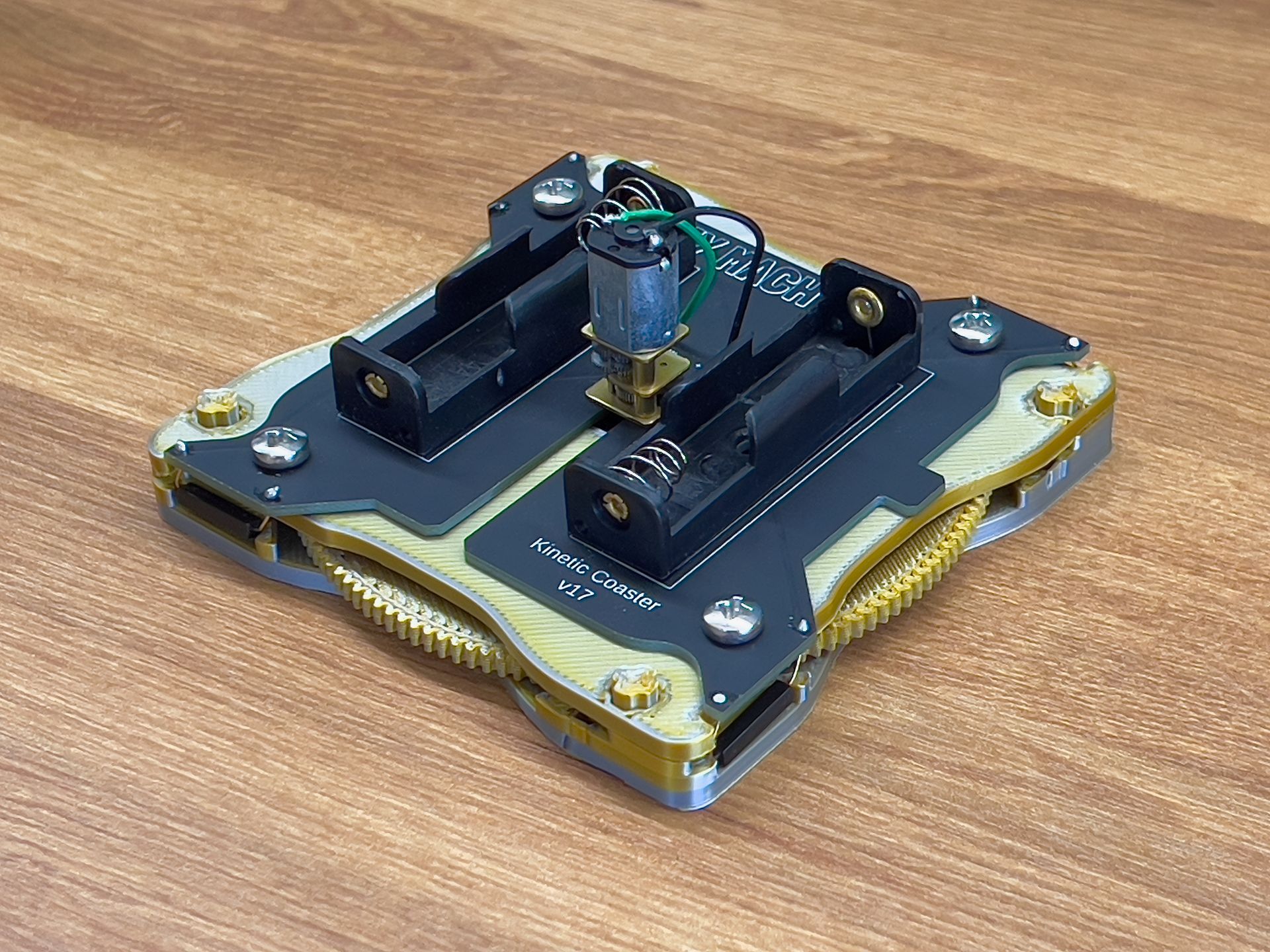 A square, brass-framed mechanical device with two battery holders and a small motor, sitting on a wooden surface.