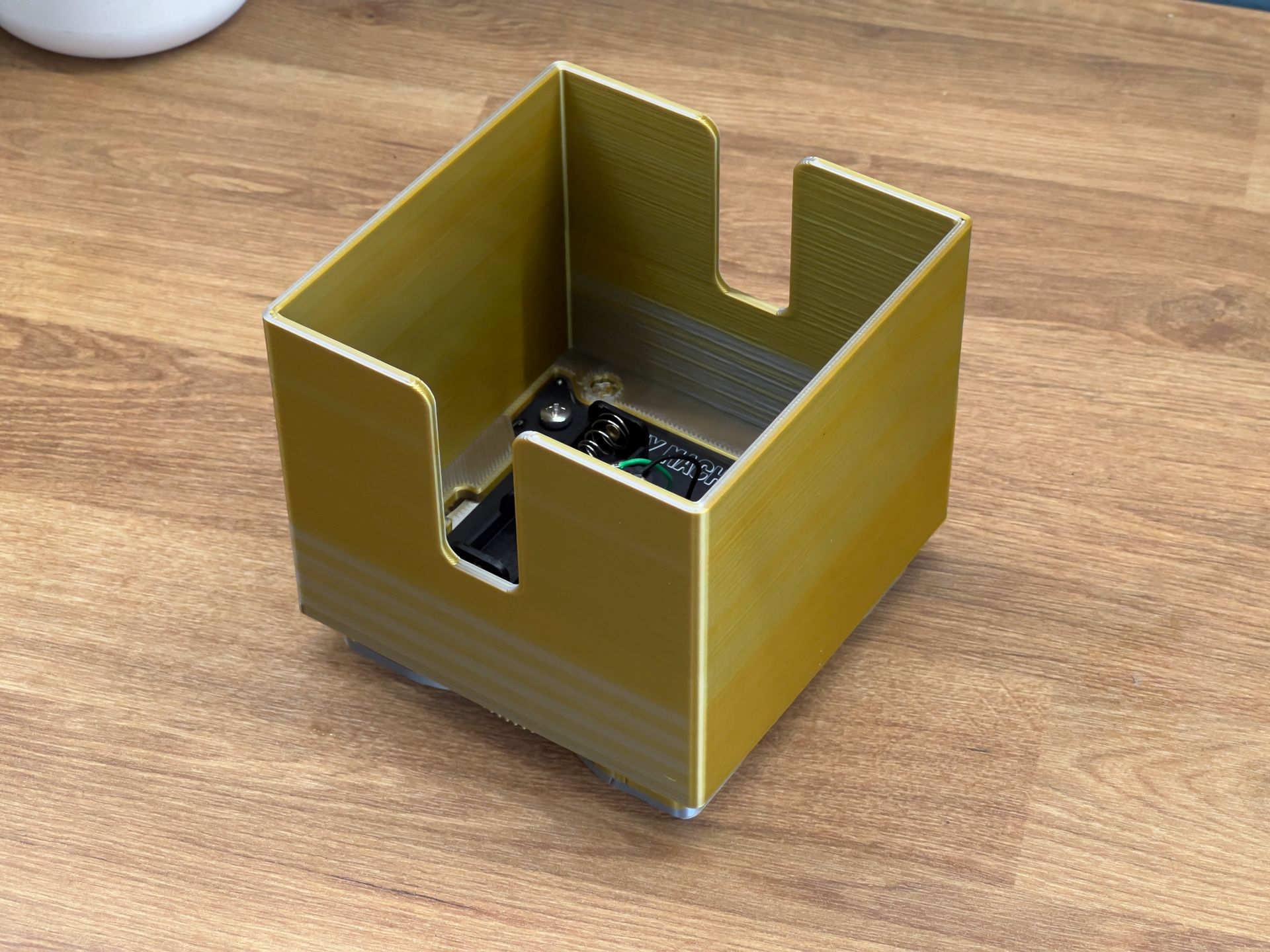 A 3D-printed, gold-colored square holder with U-shaped cutouts, sitting on a wooden surface with electronics inside.