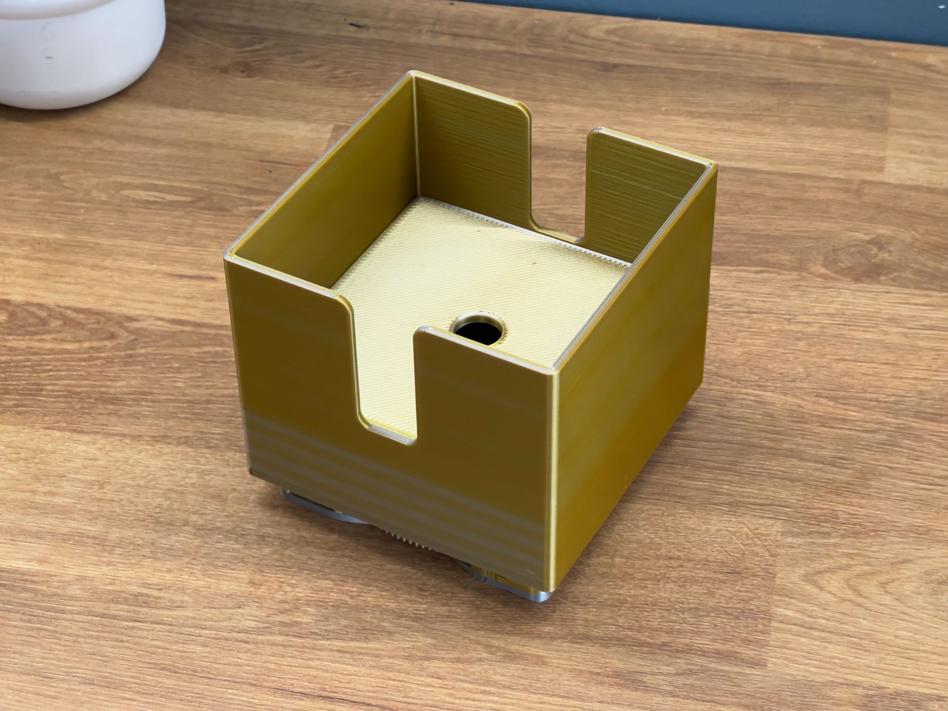 A gold 3D-printed square holder with a U-shaped cutout on the front, sitting on a wooden surface.