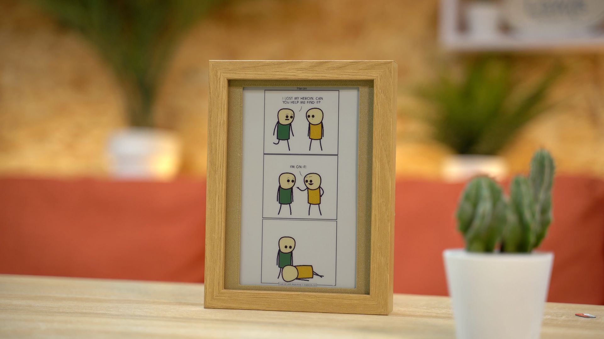 A framed cartoon on a desk featuring a three-panel comic strip with two characters interacting. A cactus sits nearby.