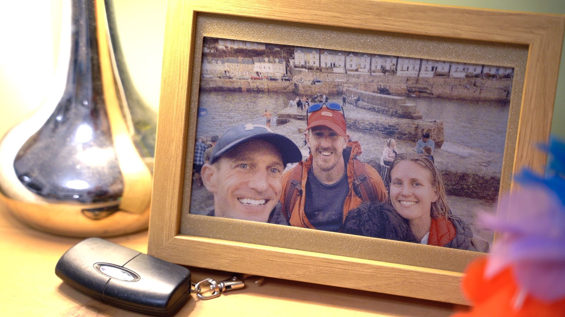 A framed photo shows three people smiling outdoors, sitting on a desk next to a shiny vase and a small black keychain.