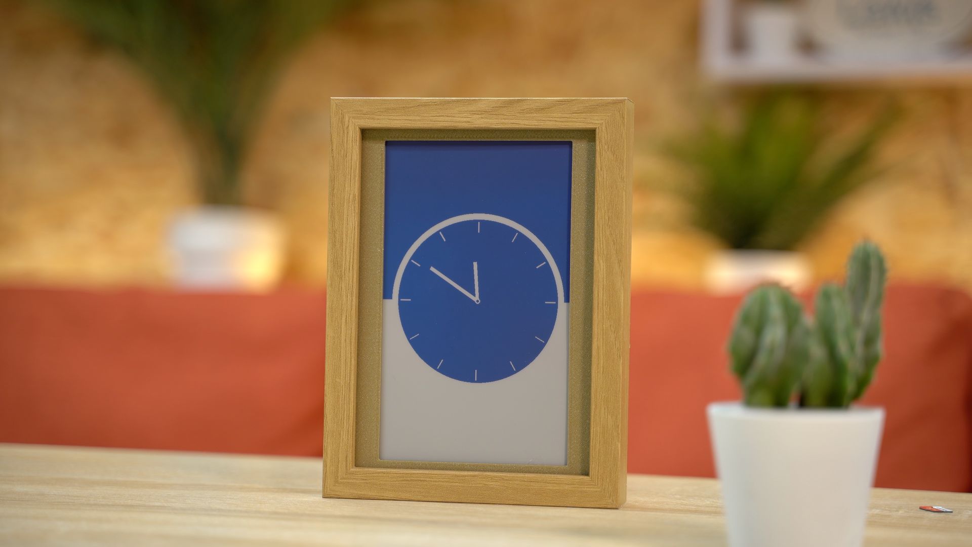 A digital clock in a wooden frame showing 10:00, sitting on a wooden table next to a small potted cactus.