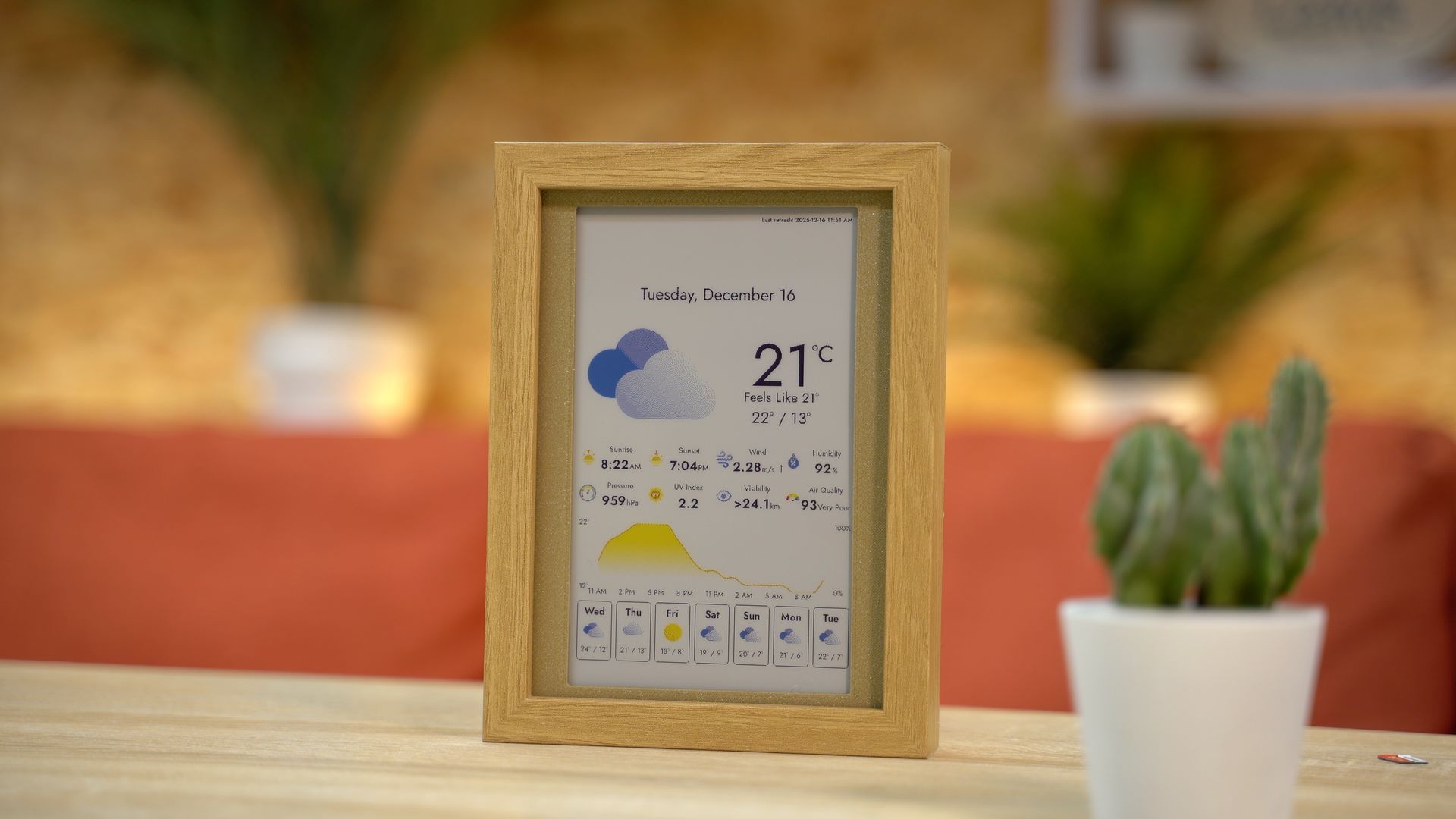 A digital weather display in a rustic wooden frame sits on a desk next to a potted cactus.