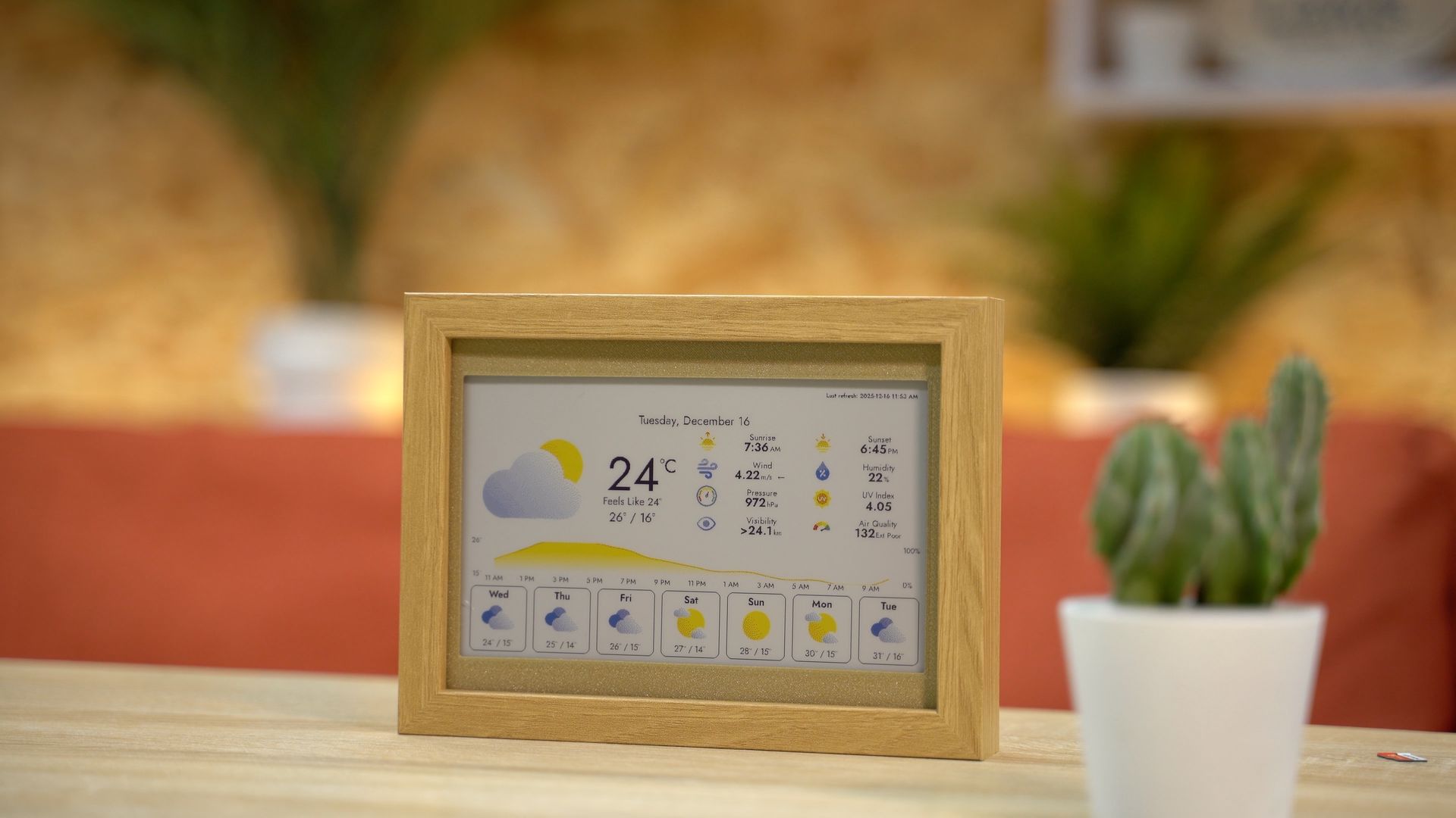 A digital weather display in a wooden frame sits on a wooden surface next to a small potted cactus.