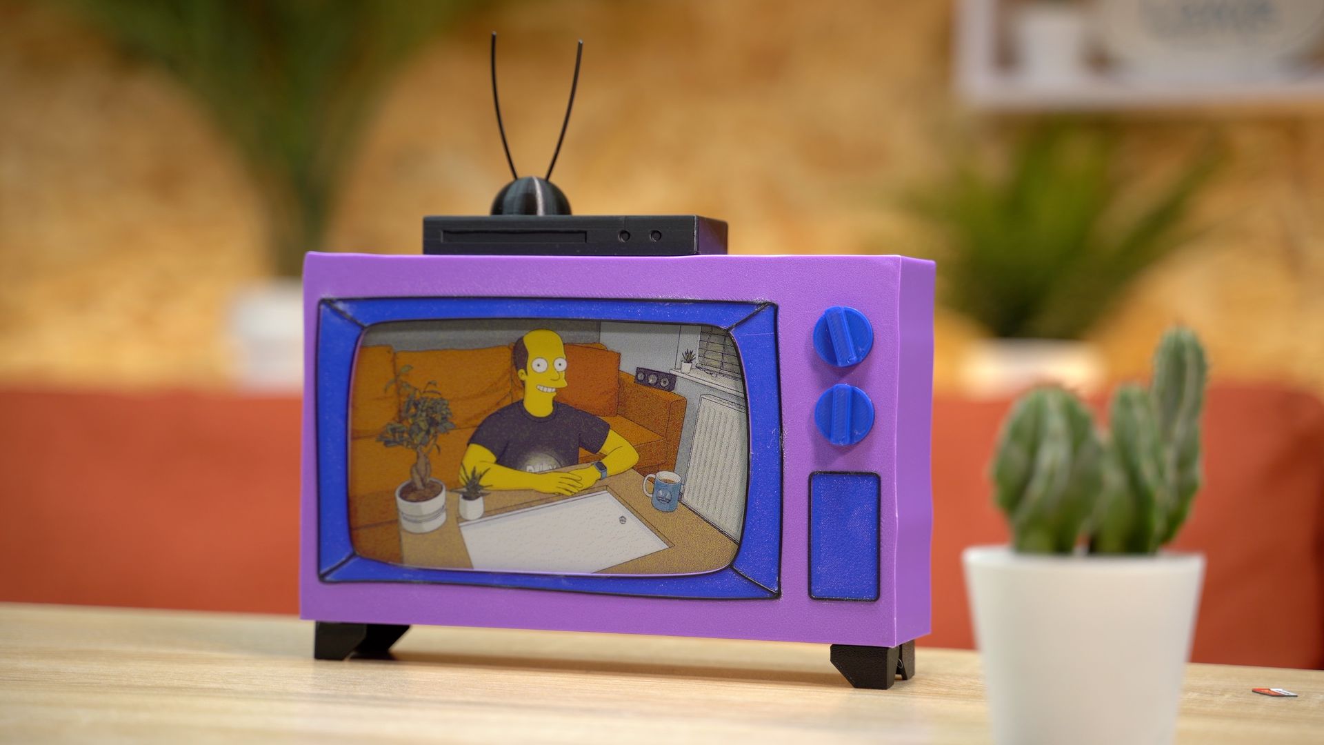 A small, purple, toy television with rabbit ears displays an animated scene of a person sitting at a desk indoors.