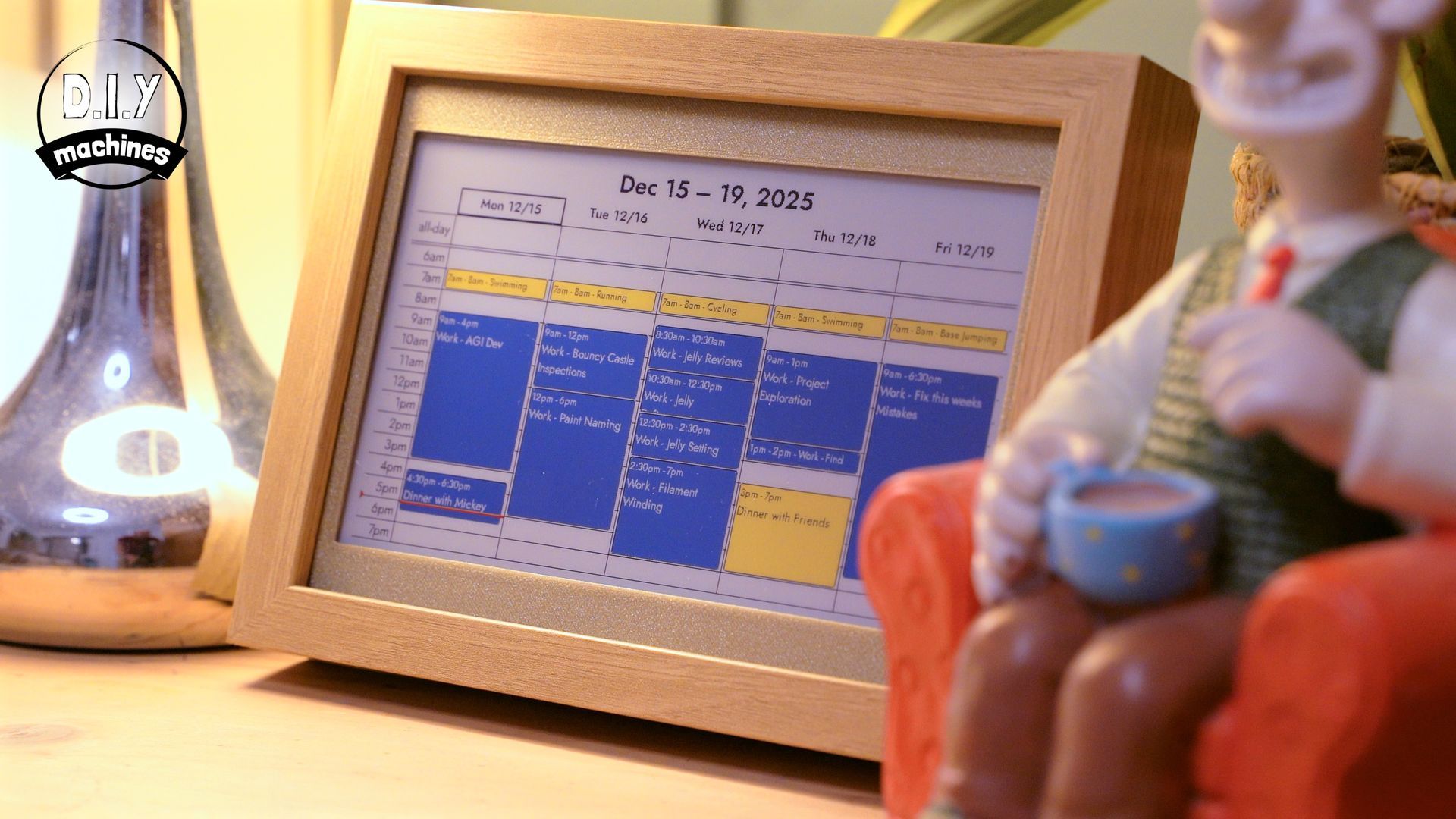 A wooden-framed digital calendar sits on a desk next to a figurine of a man in an armchair holding a mug.