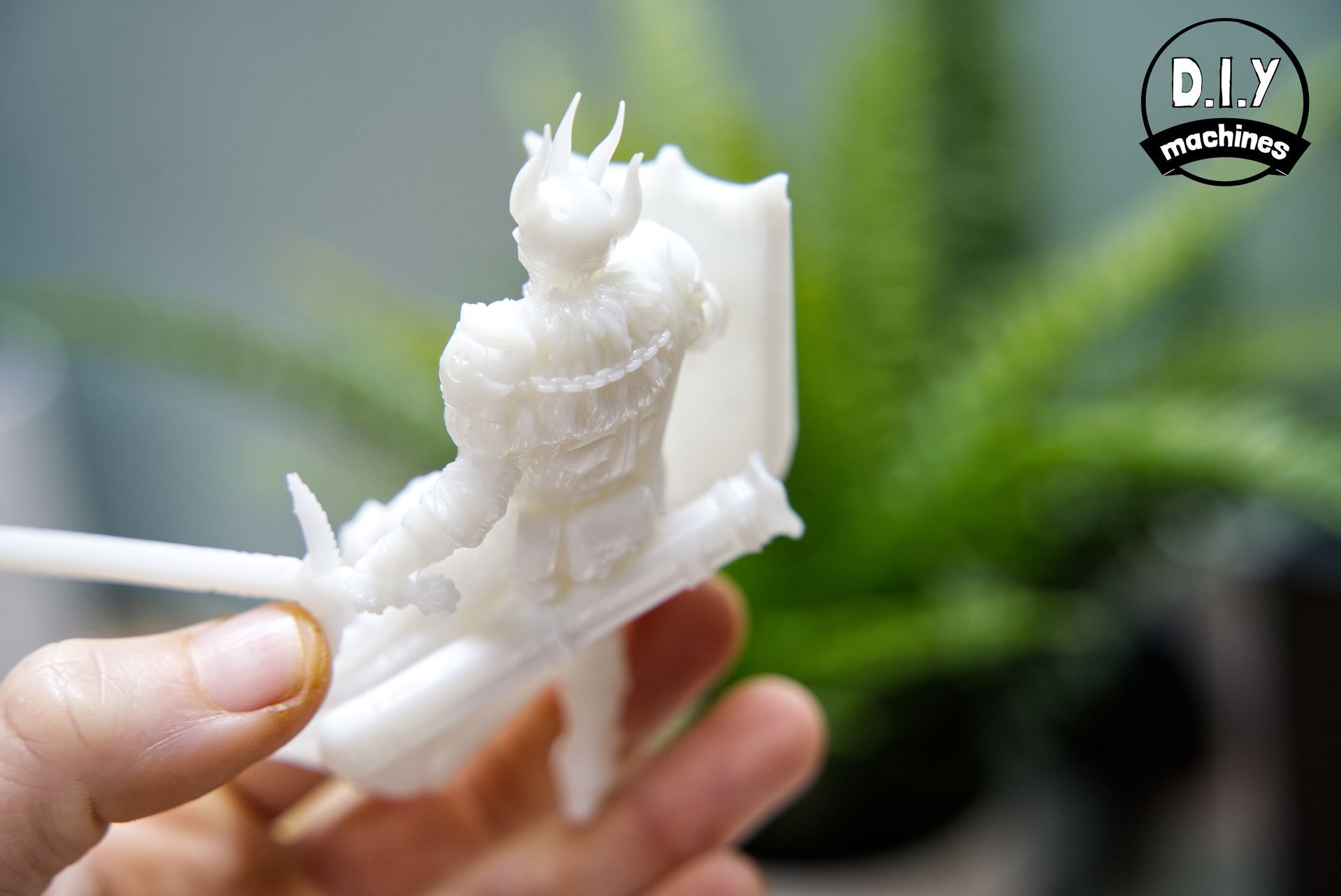 A hand holds an unpainted white 3D-printed miniature figure of a fantasy knight in armor, with a plant in the background.