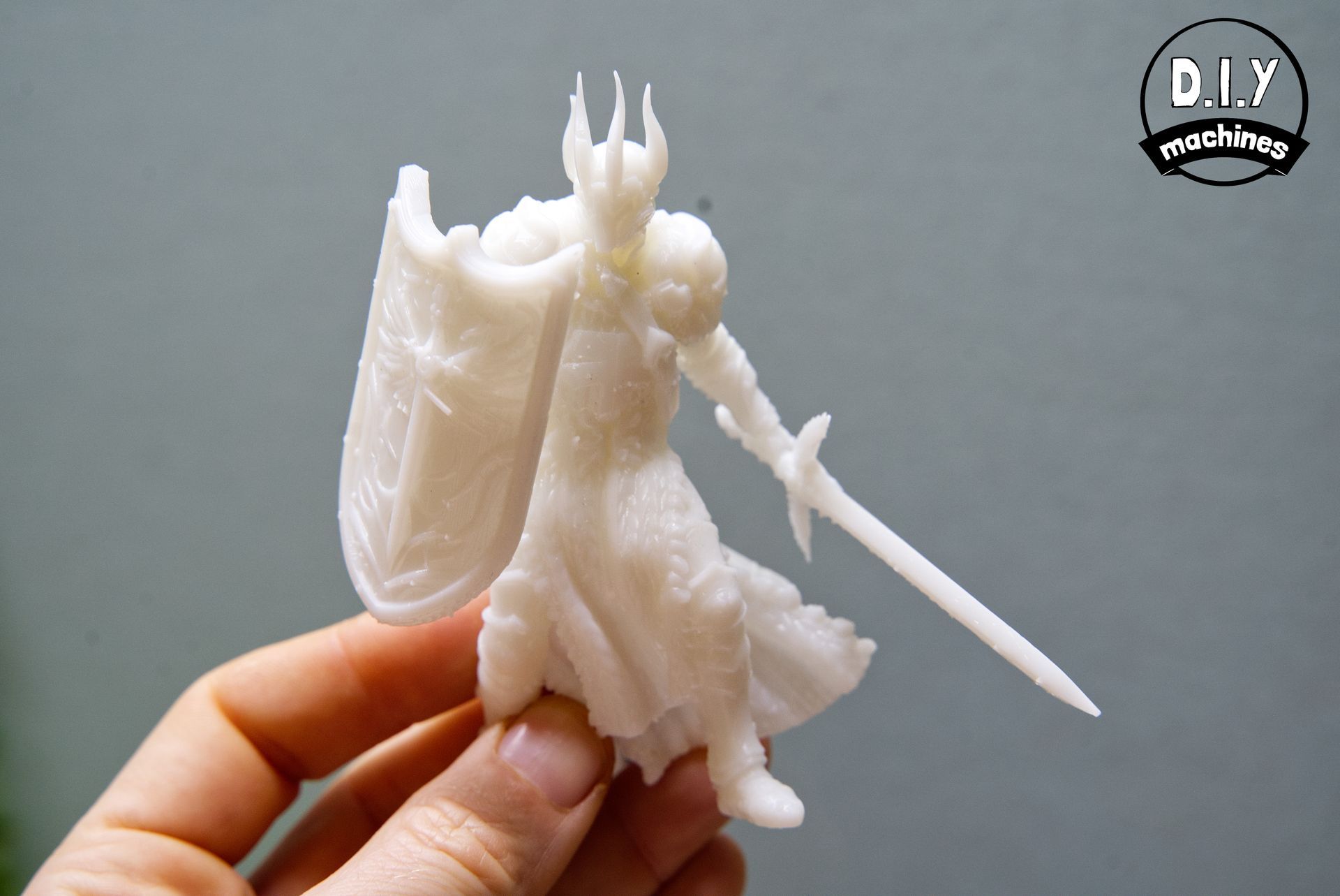 A person holds a white 3D-printed knight figurine with a crown, shield, and sword against a grey background.