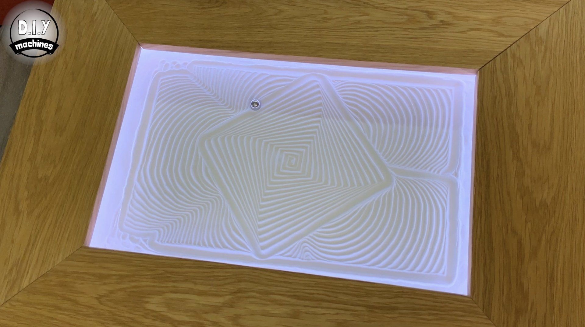 A motorized metal ball draws intricate, geometric patterns in white sand within a wooden frame on a table.