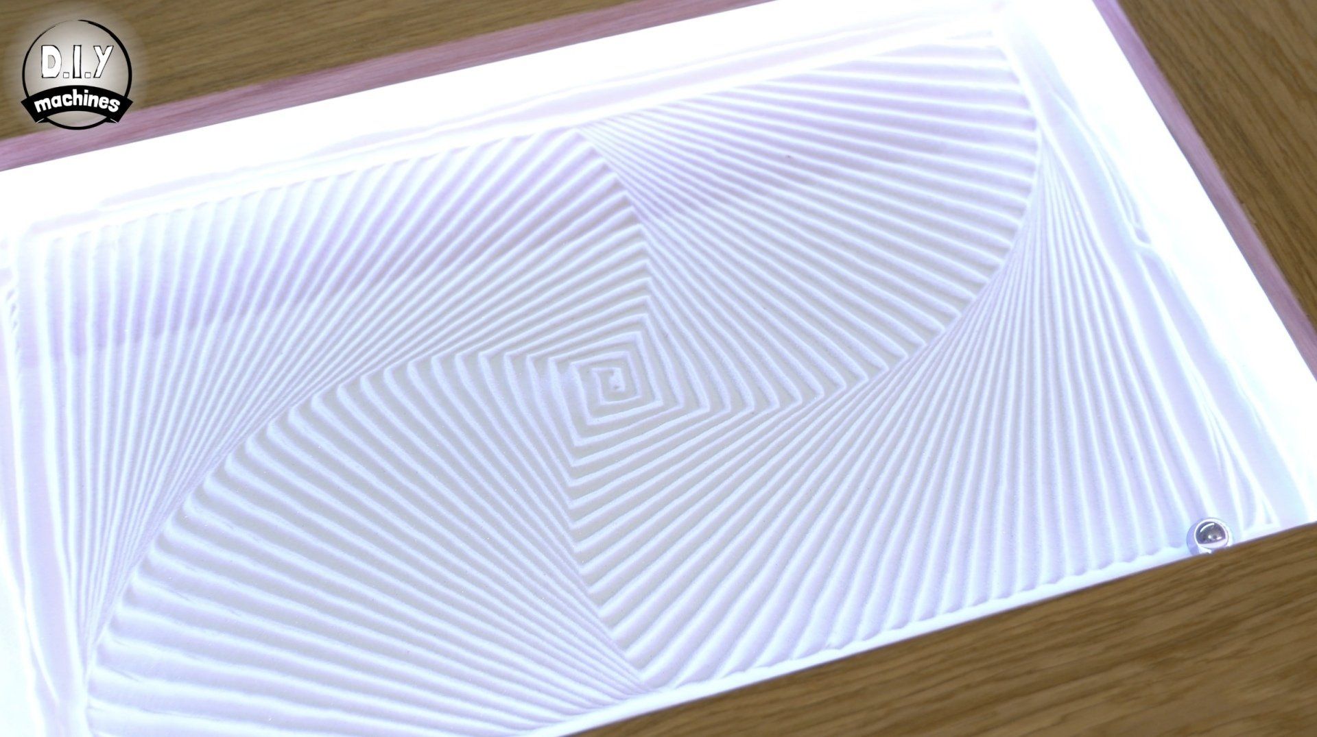 A kinetic sand table displays an intricate, spiraling white geometric pattern on a light background.