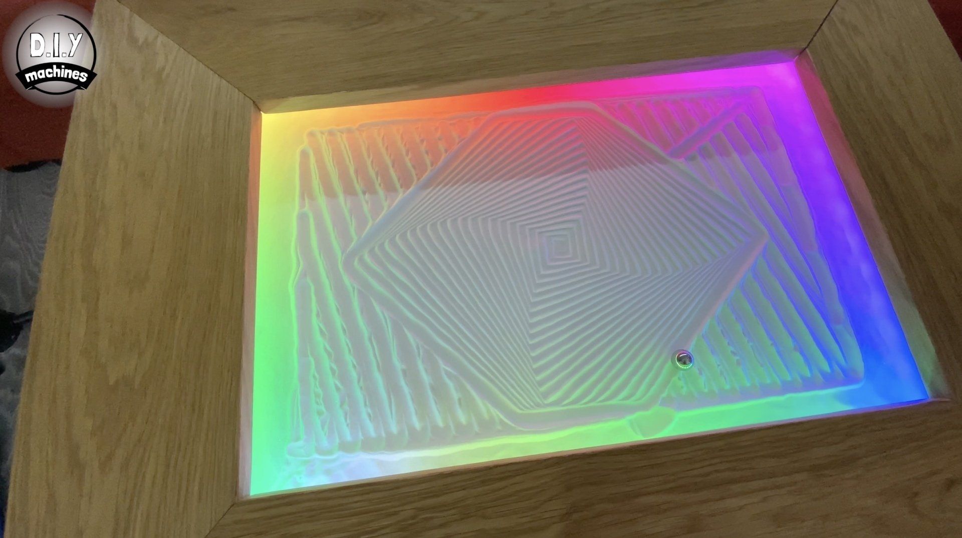A wooden-framed sand table illuminated with rainbow LED lights, displaying a geometric pattern traced in the sand.