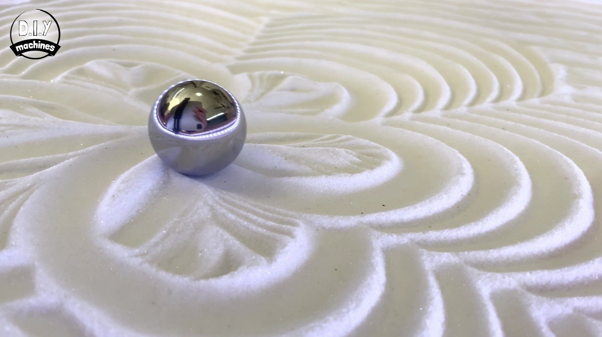 A metallic sphere resting on a bed of white sand, featuring intricate, concentric ripple patterns carved into the surface.