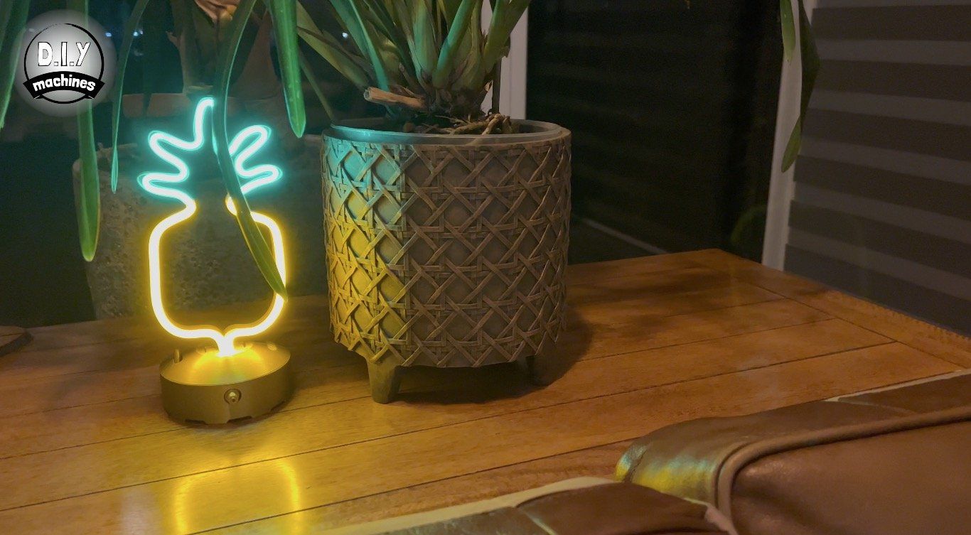 A glowing, two-tone neon pineapple lamp sits on a wooden surface next to a textured, grey-patterned plant pot.