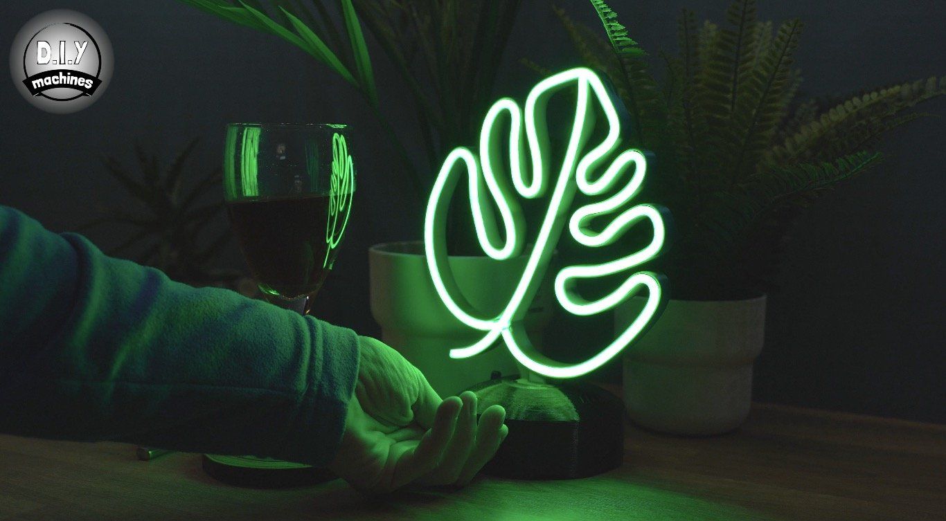 A person's hand reaches toward a glowing neon monstera leaf sign on a dark table next to a glass of wine and house plants.