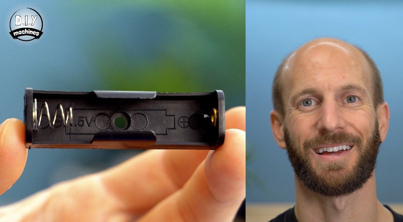 A person holds a black plastic AA battery holder next to their face, looking at the camera with a smile.