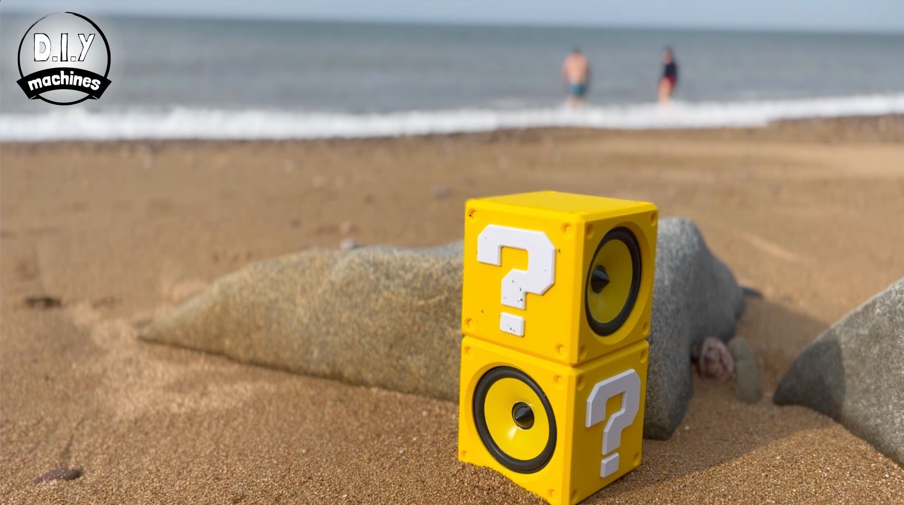 Two stacked yellow Mario question mark blocks serving as speakers sit on a sandy beach near the ocean.
