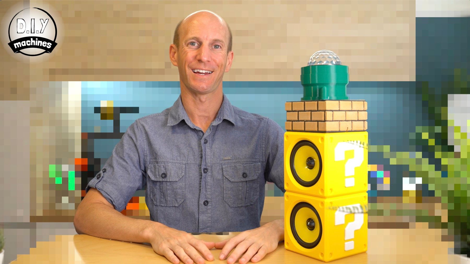 A smiling person sits at a desk next to a tower of two yellow Mario-style question blocks topped with a green brick.