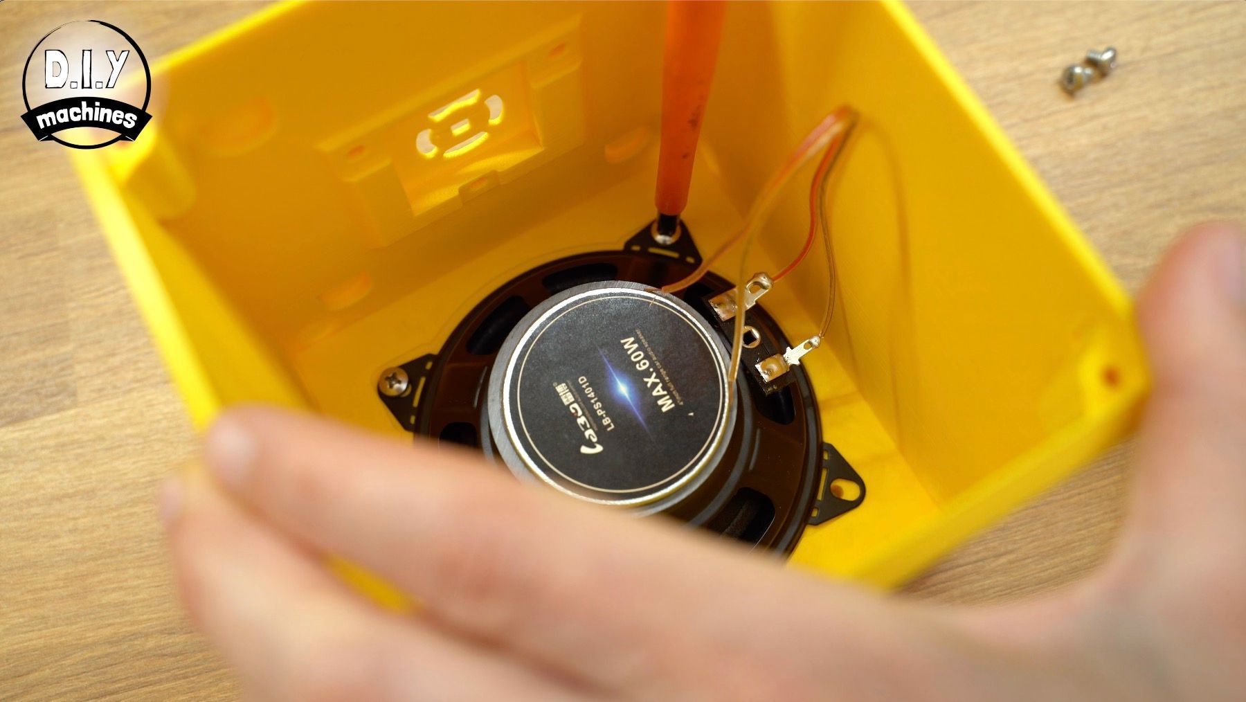 A speaker is being installed inside a bright yellow, open-top plastic enclosure.
