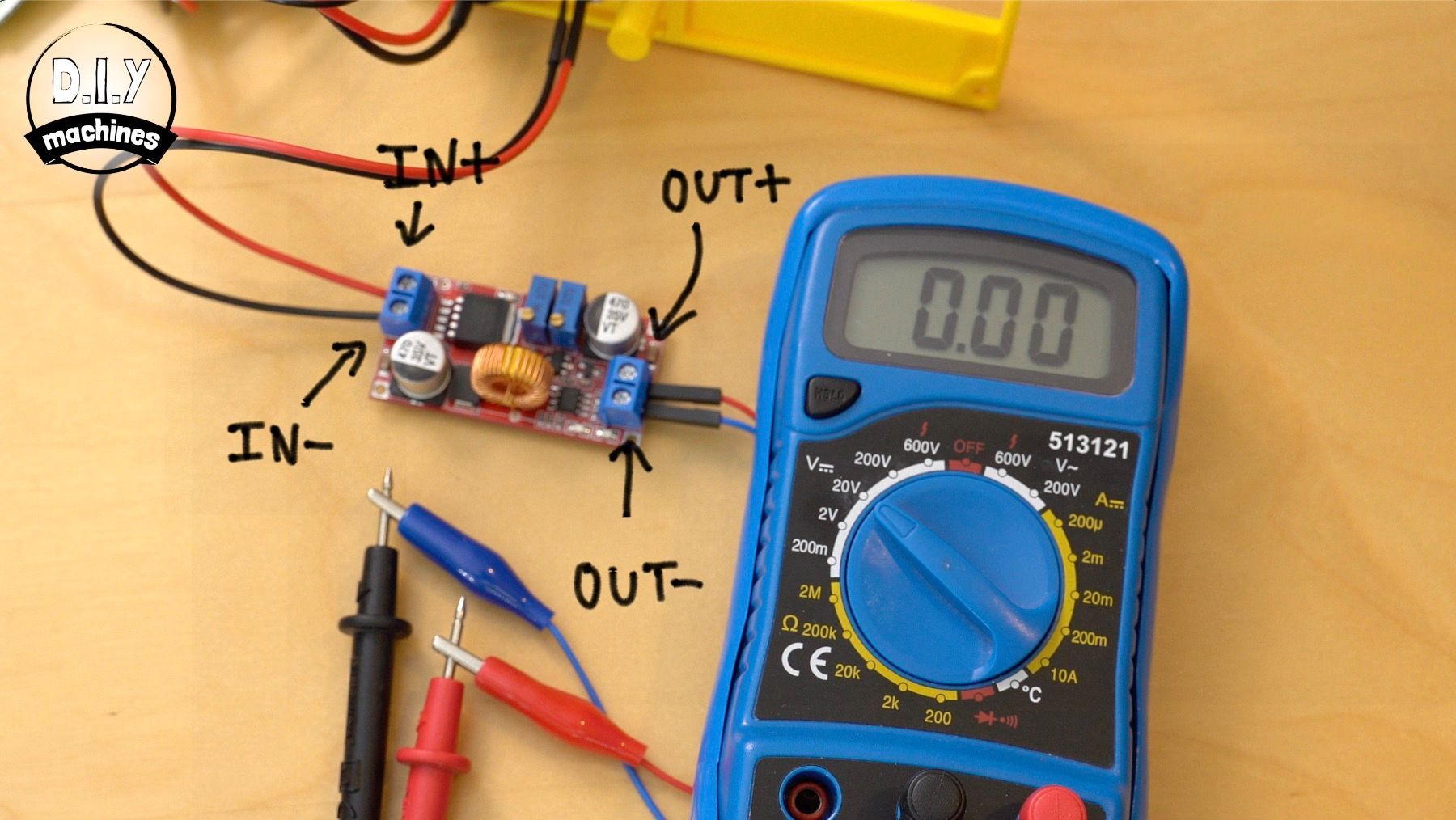 A buck converter module connected to a blue digital multimeter, with wires labeled IN+, IN-, OUT+, and OUT- on a table.