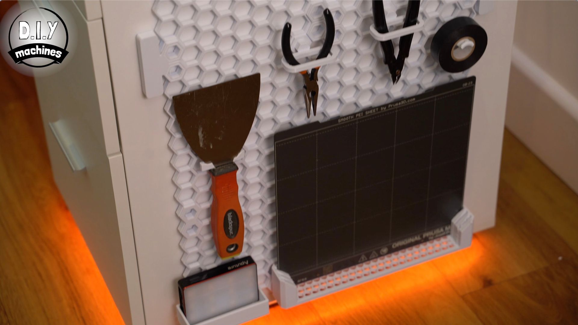 A white honeycomb pegboard attached to a furniture side holds tools, a putty knife, a build plate, and electrical tape.