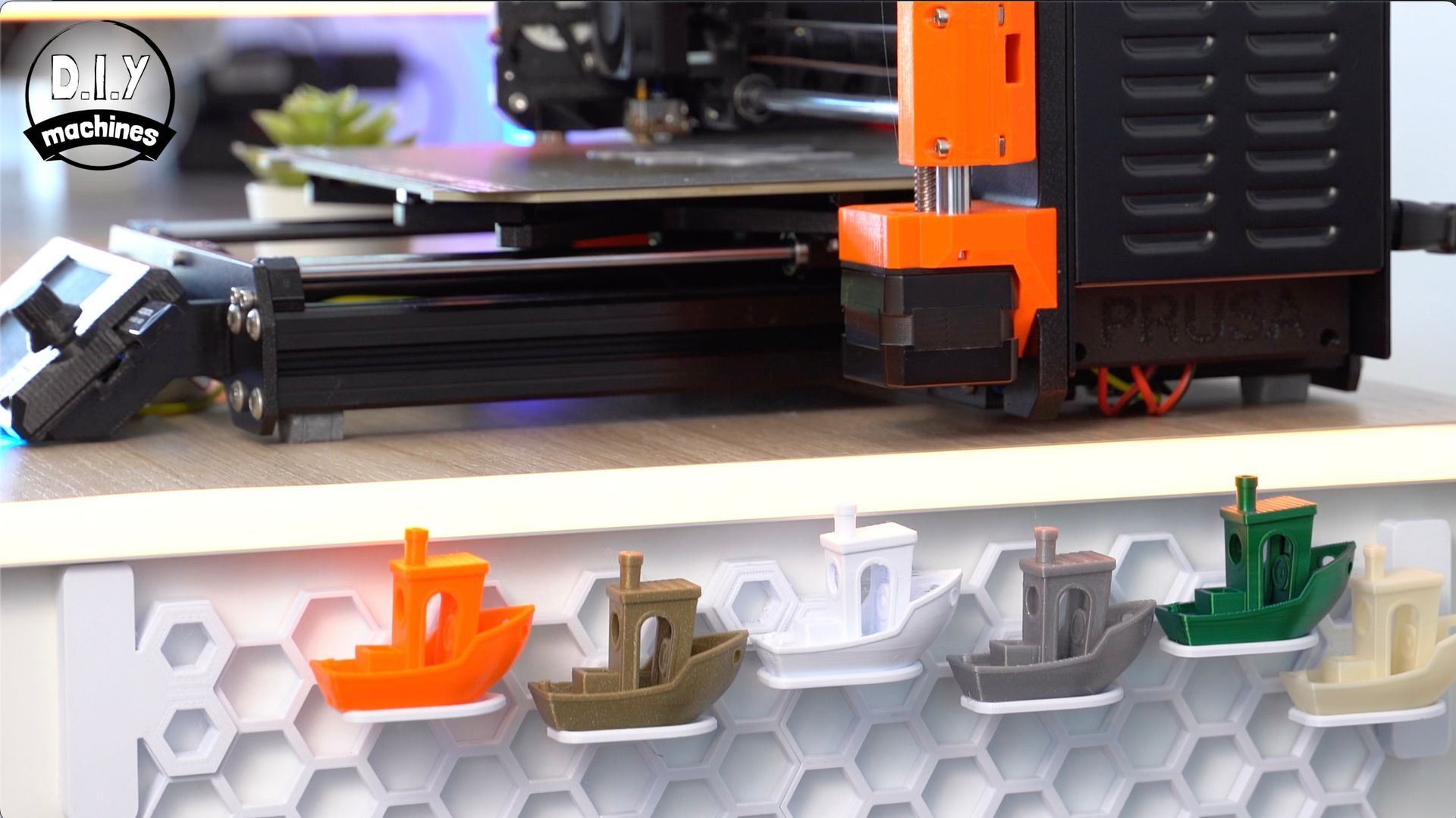 A 3D printer sits above a display shelf featuring six small, multicolored toy boats mounted on a hexagonal wall panel.