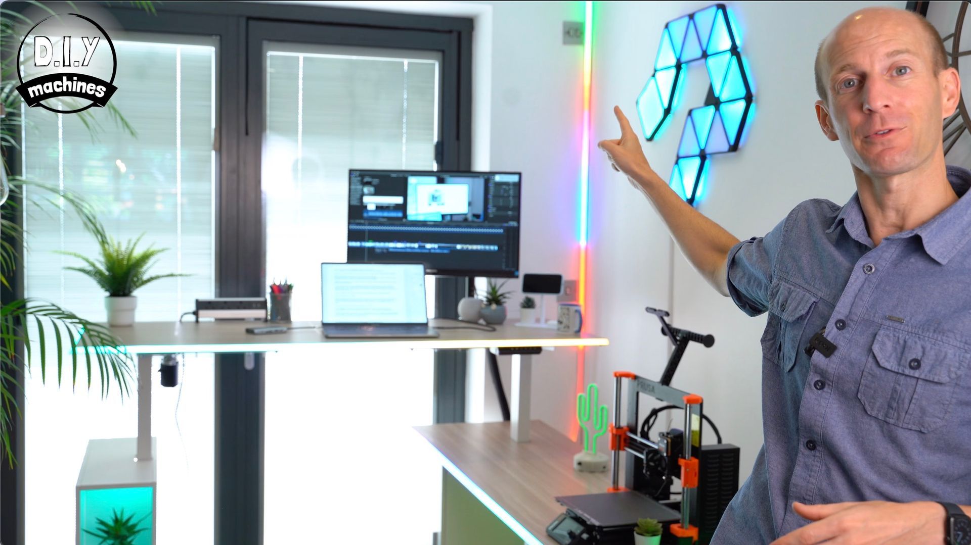 A person points toward a wall-mounted light display in a home office with a standing desk and 3D printer.