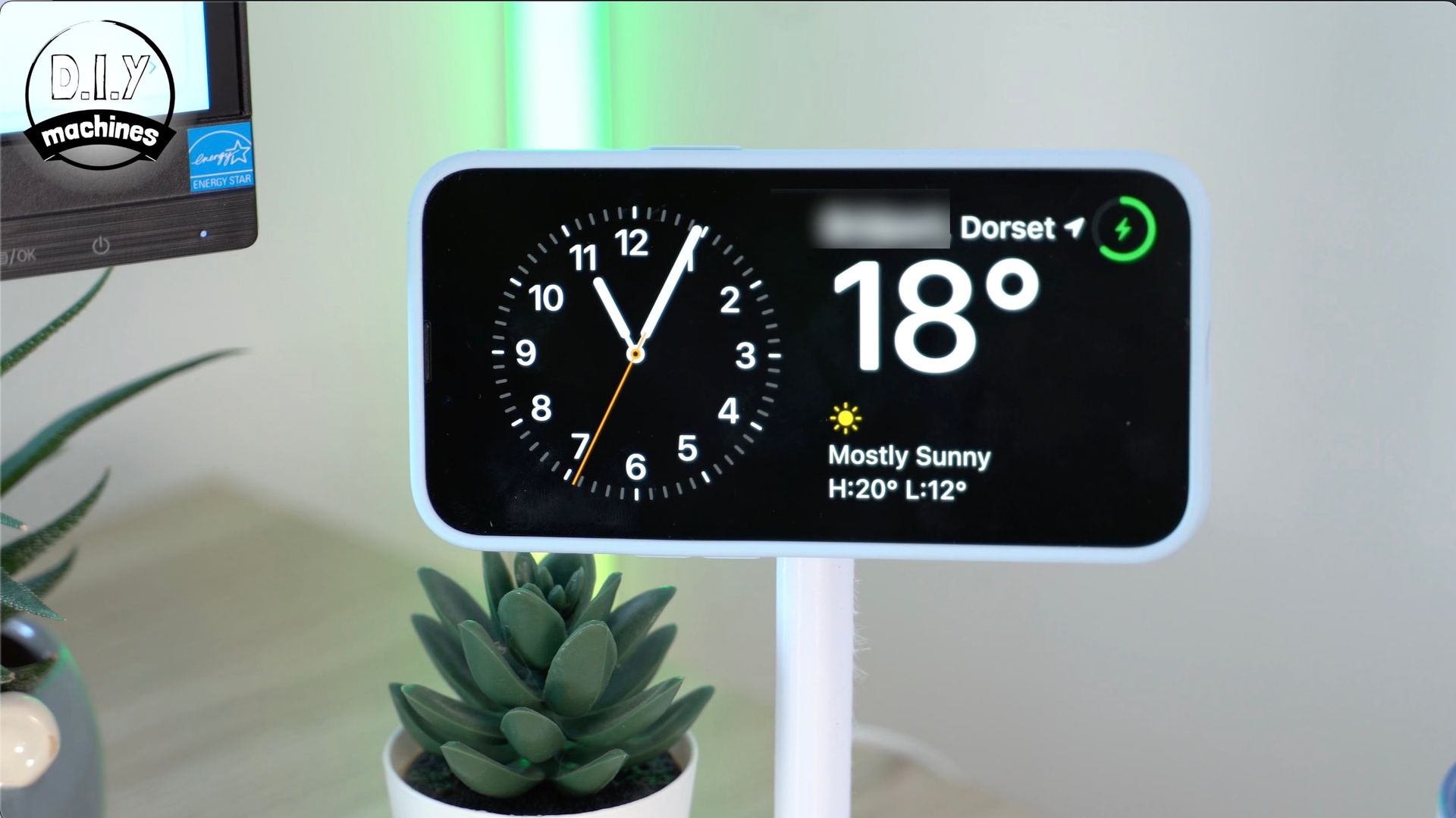 A smartphone on a stand displays a digital clock widget with weather info for Dorset, showing 18 degrees and mostly sunny.