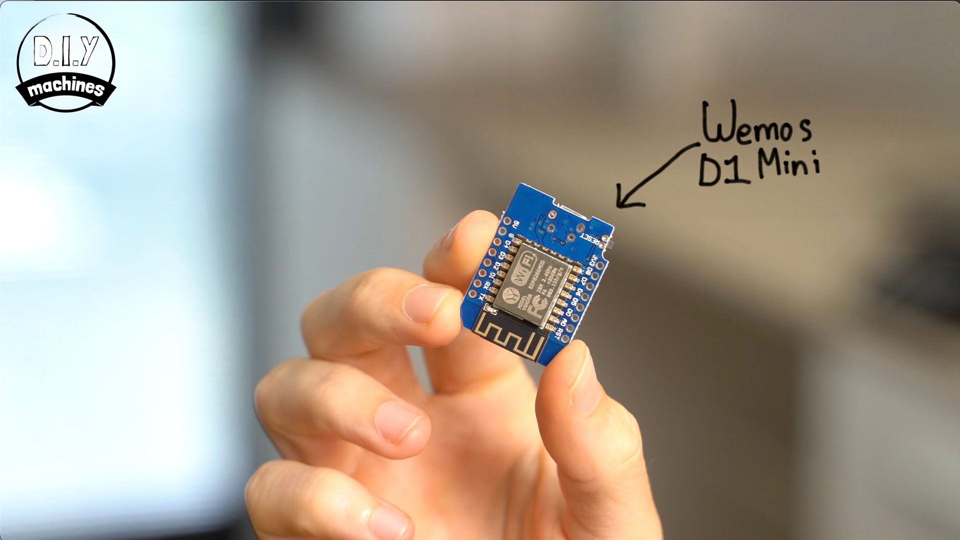A person’s hand holds a small, blue Wemos D1 Mini microcontroller board against a blurred indoor background.