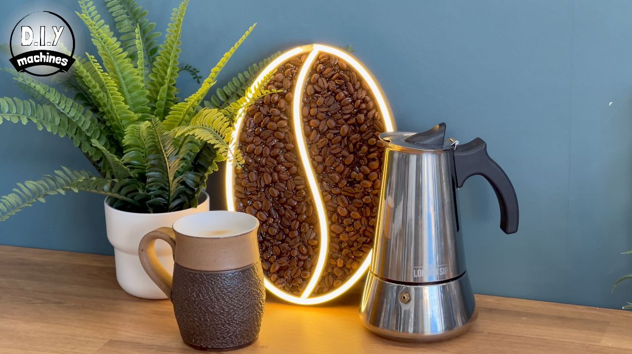 A decorative glowing coffee bean sign sits on a wooden surface next to a fern in a white pot, a mug, and a steel moka pot.