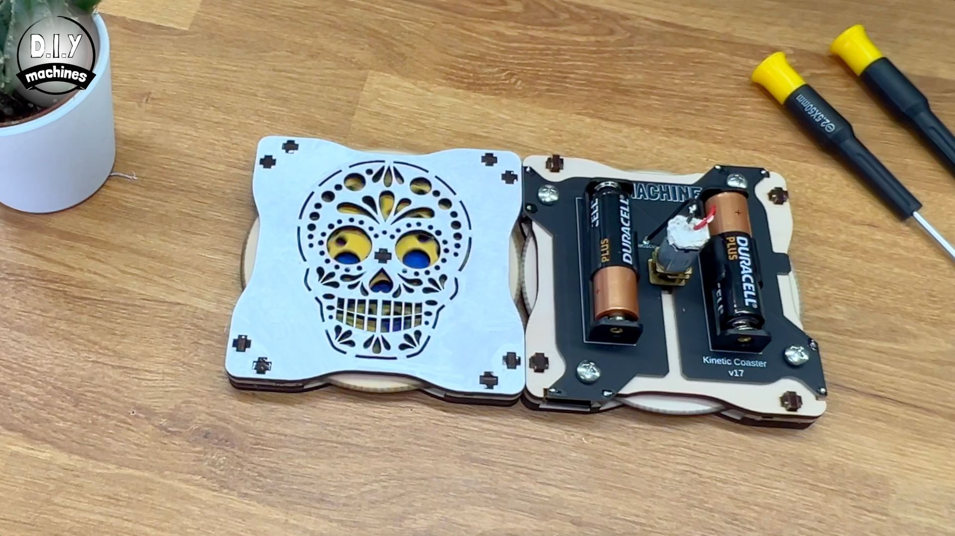 A two-part, laser-cut wooden assembly features a sugar skull design on one side and two batteries on the other.