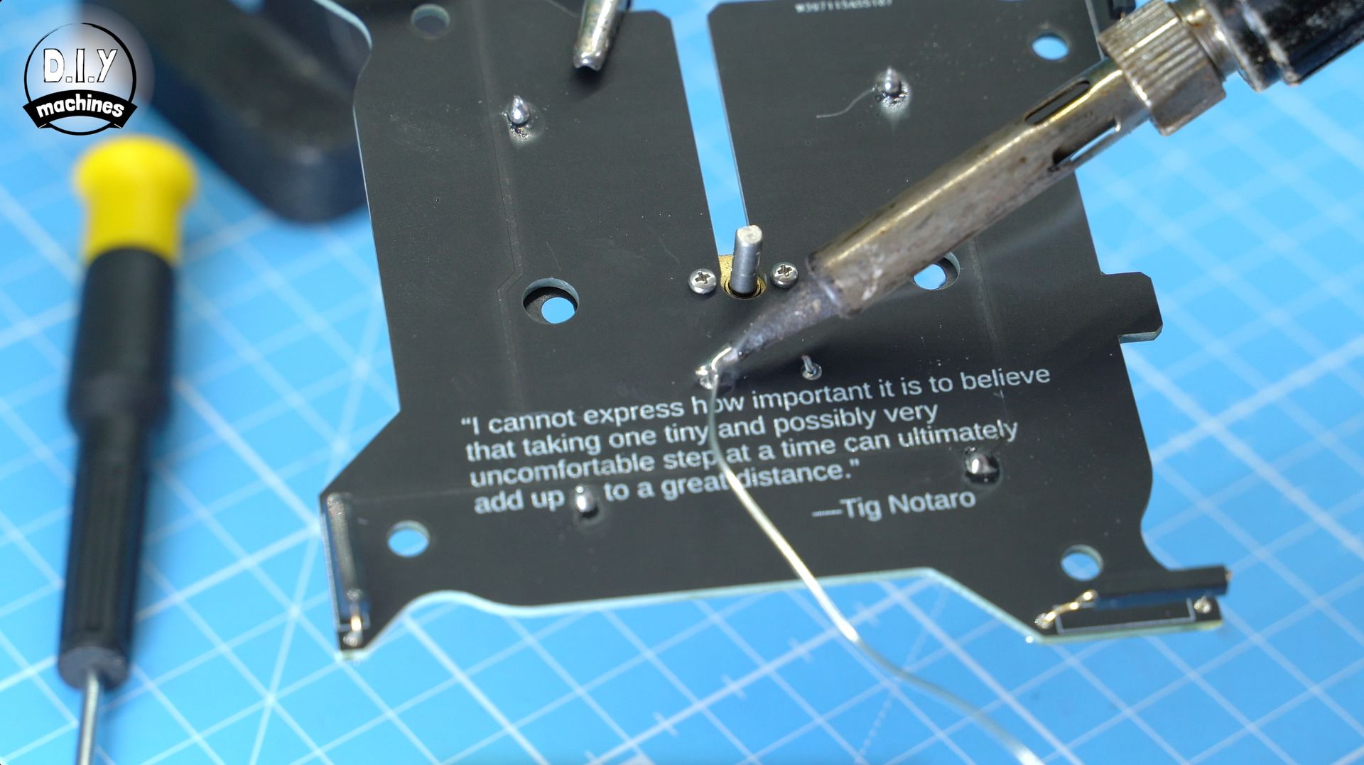 A soldering iron tip melts wire onto a black circuit board printed with a motivational quote on a blue grid mat.