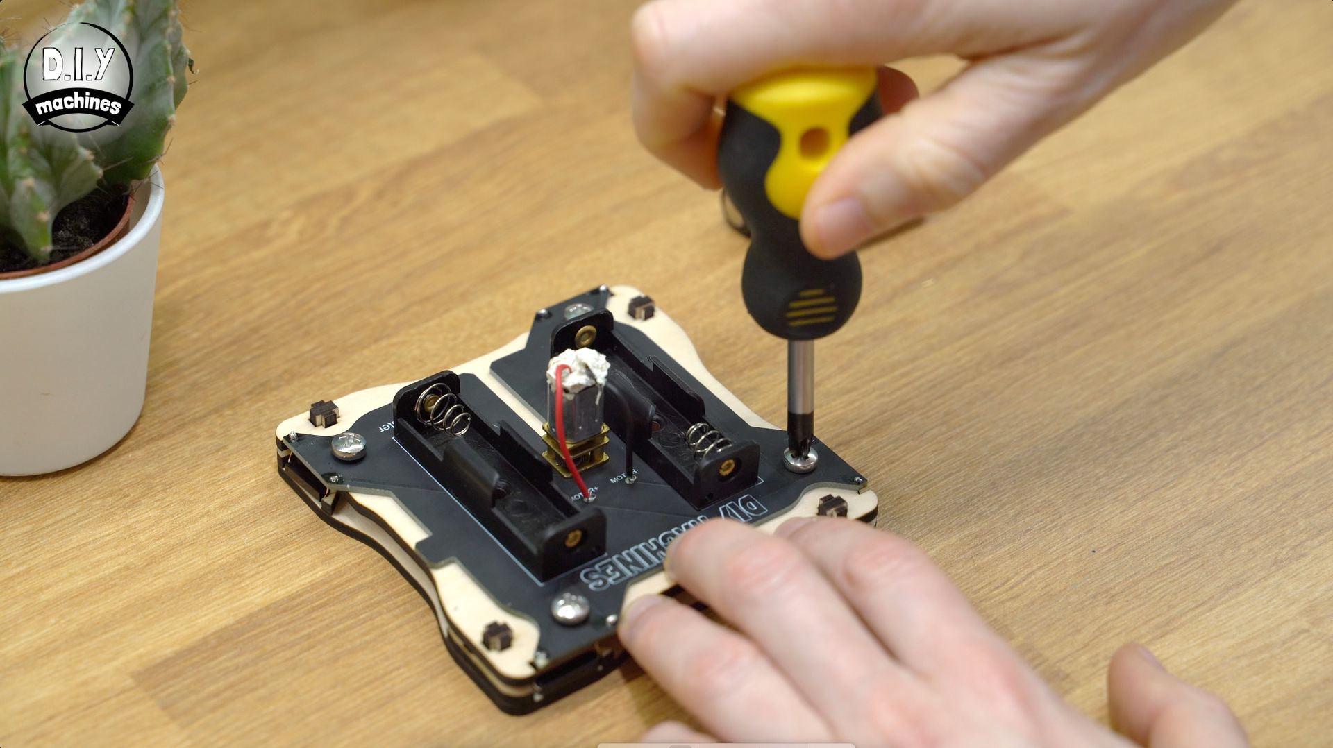 A person uses a yellow-handled screwdriver to secure a screw on an electronic circuit board with two battery holders.