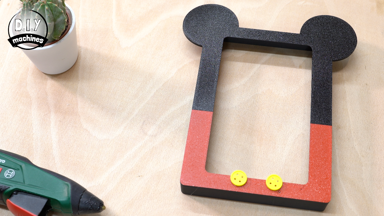A DIY Mickey Mouse-themed frame made of black and red foam with two yellow buttons on a wooden table next to a glue gun.