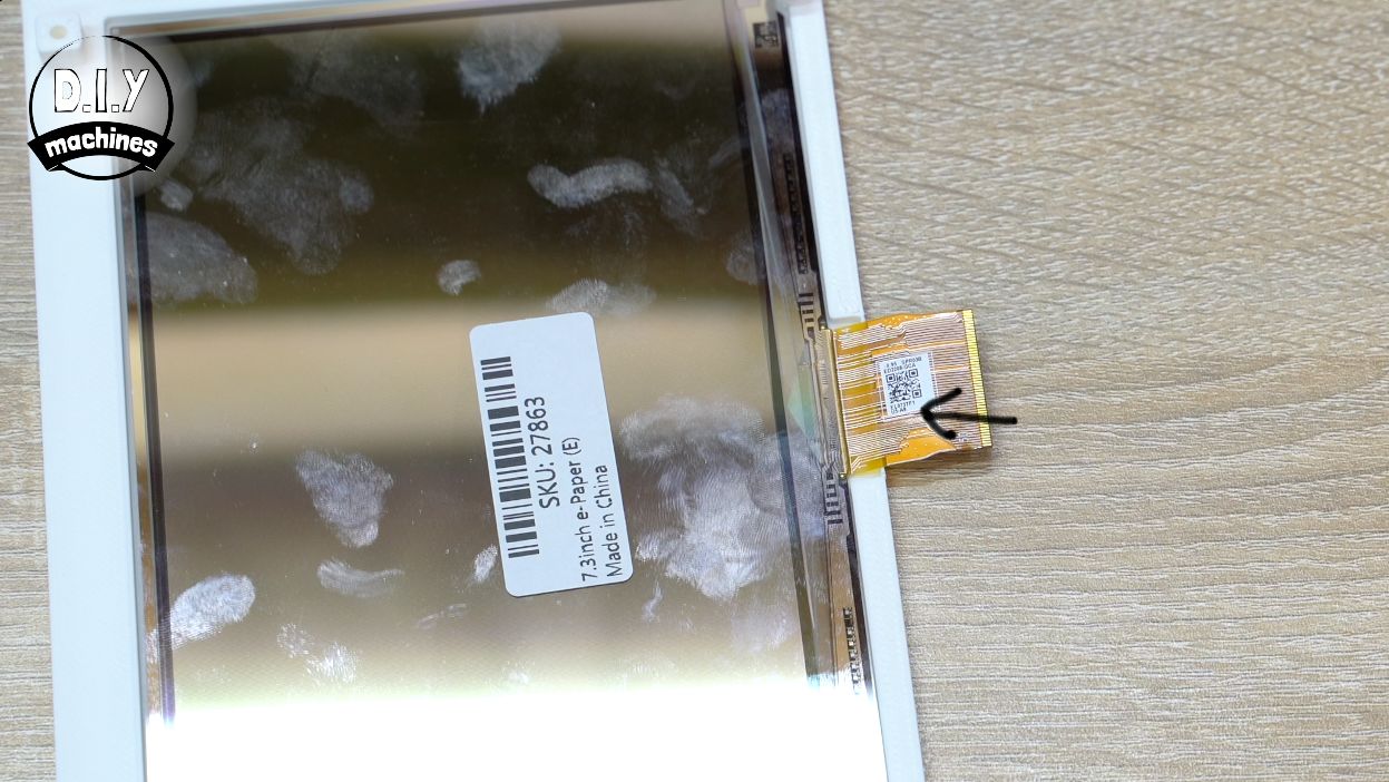A rectangular screen component on a table with a small golden ribbon cable connector highlighted by a black arrow.