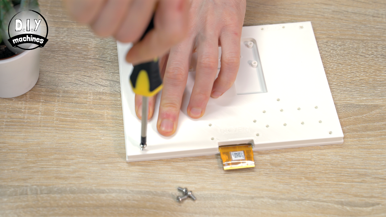 A person uses a yellow and black screwdriver to fasten a small screw into a white panel on a wooden table.