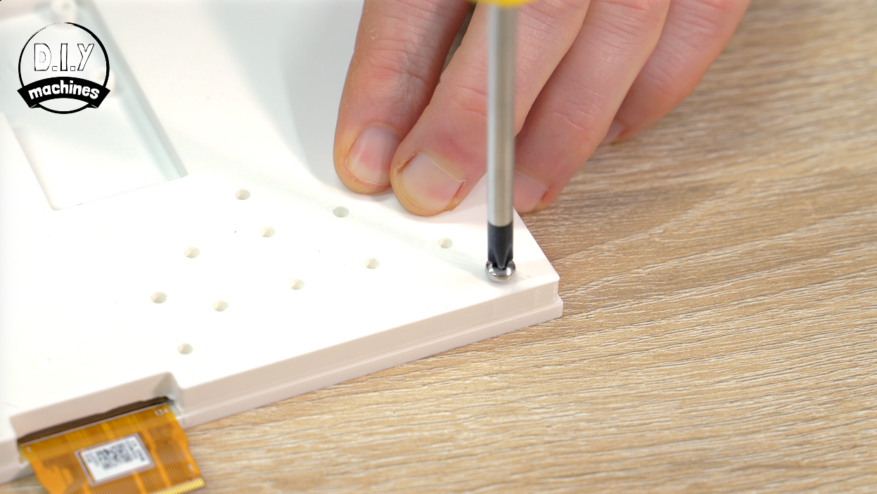 A hand uses a screwdriver to tighten a screw into a white plastic component on a wooden work surface.