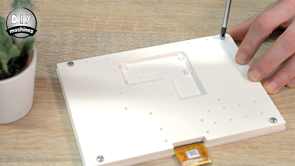 A hand uses a screwdriver on a white rectangular device component with a ribbon cable, placed on a wooden surface.