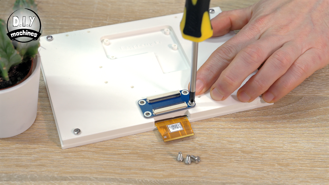 A hand uses a screwdriver to mount a small blue electronic component onto a white surface with loose screws nearby.