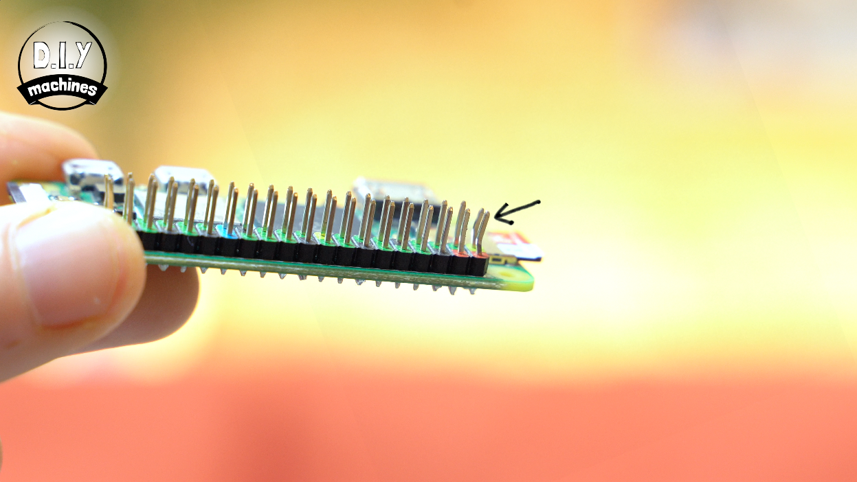 A hand holds a green Raspberry Pi Zero board with a row of bent header pins, highlighted by a small black arrow.