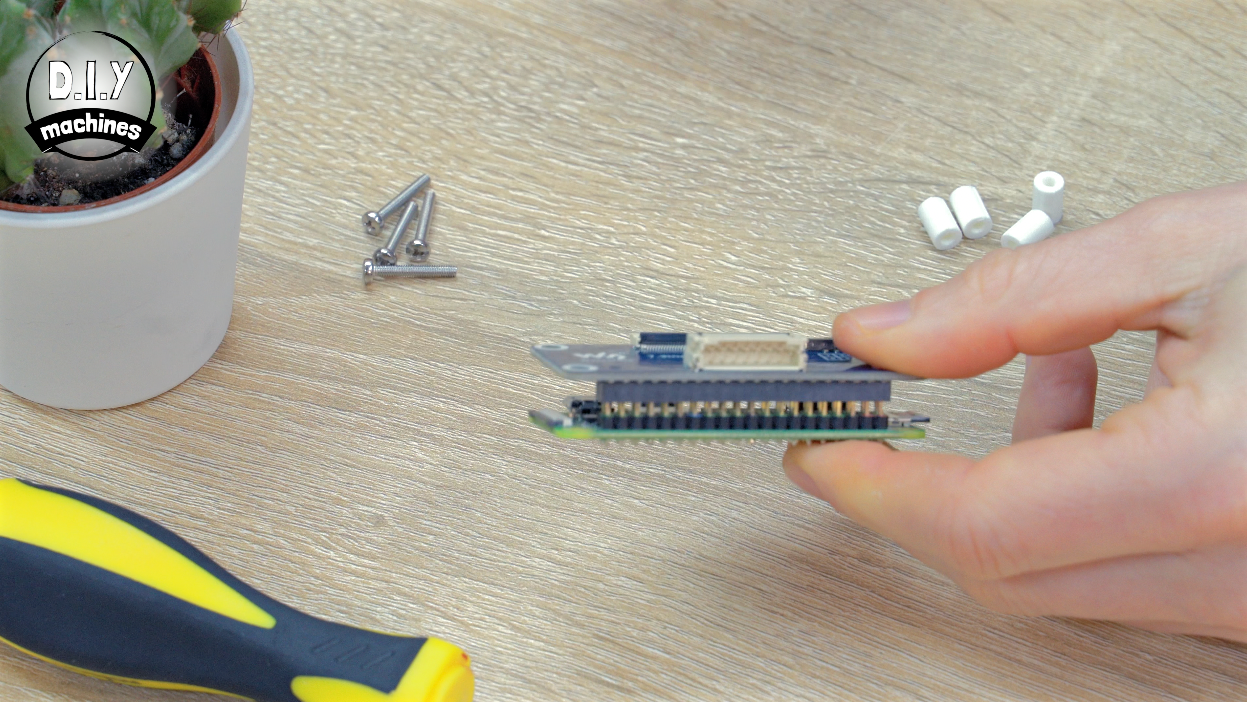 A hand aligns a small electronics module over a header on a circuit board, with screws and spacers nearby on a table.