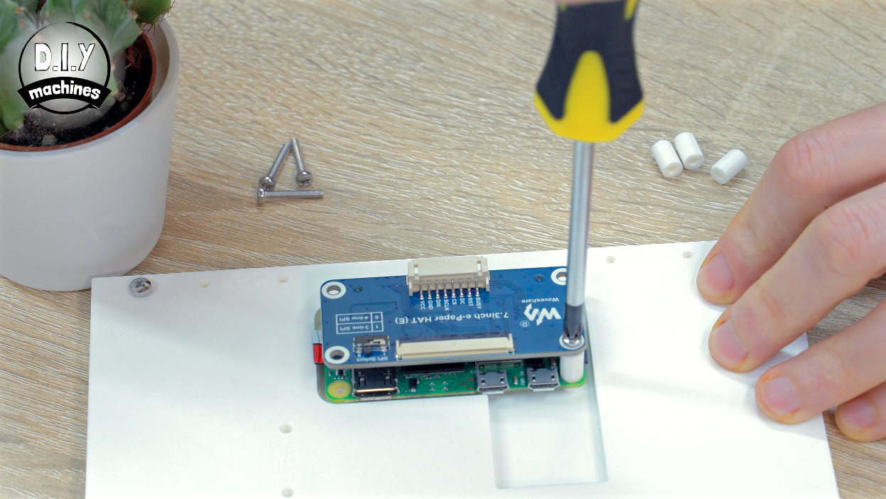 A person uses a yellow-handled screwdriver to mount a small circuit board onto a white surface with plastic spacers.