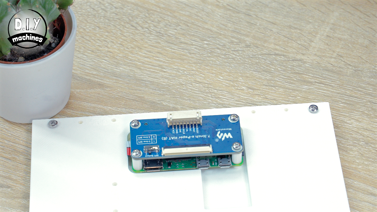 A blue printed circuit board mounted on a white plastic sheet next to a potted succulent on a wooden table.