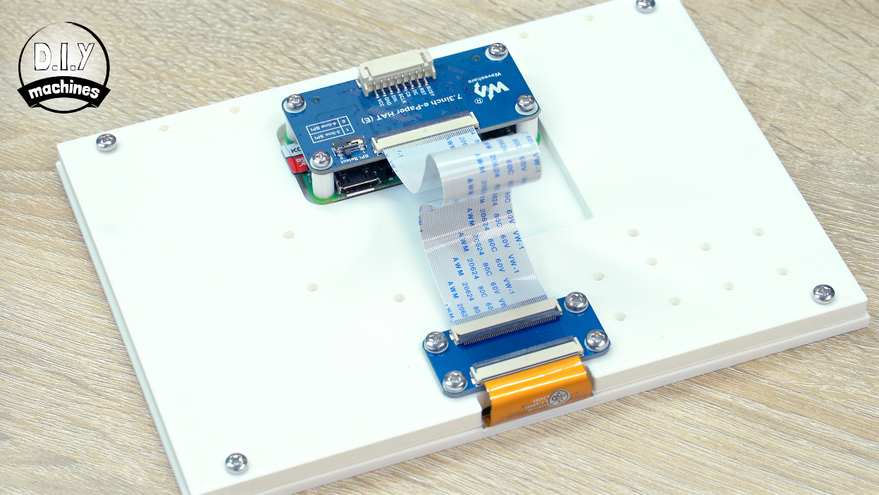 A white display screen mounted on a flat panel, connected to a blue electronic circuit board by a flat ribbon cable.